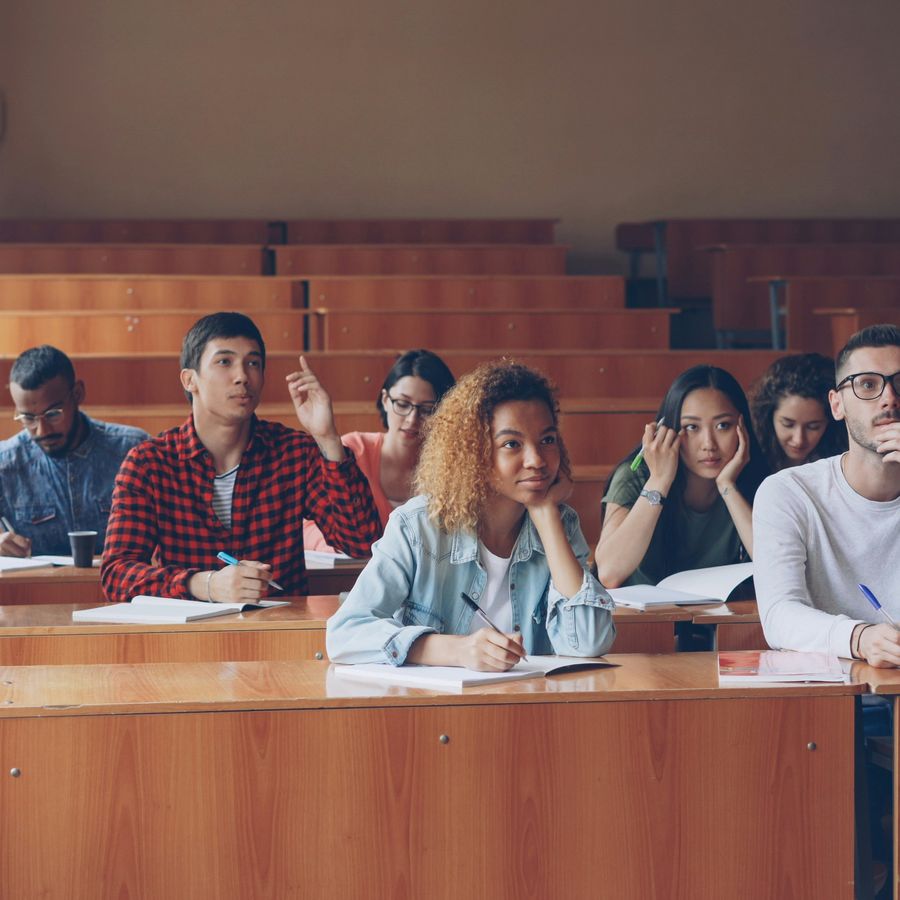 Students seated at tiered desks in a university lecture hall, taking notes, one raising a hand.