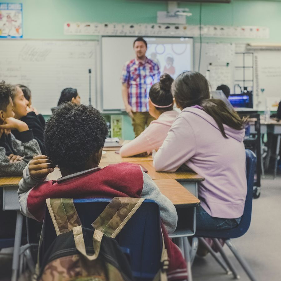 A middle school class seated at tables, watching a teacher present at the front of the room.